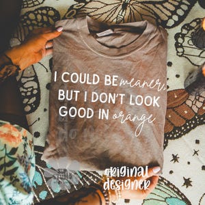 May include: A brown t-shirt with the text "I COULD BE meaner, BUT I DON'T LOOK GOOD IN orange" in white. The shirt is folded and held by hands, resting on a patterned surface with butterfly designs. The words "original designer" are also visible.