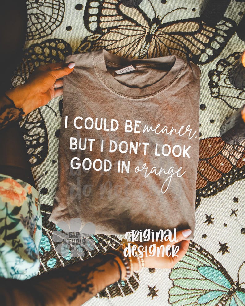 May include: A brown t-shirt with the text "I COULD BE meaner, BUT I DON'T LOOK GOOD IN orange" in white. The shirt is folded and held by hands, resting on a patterned surface with butterfly designs. The words "original designer" are also visible.