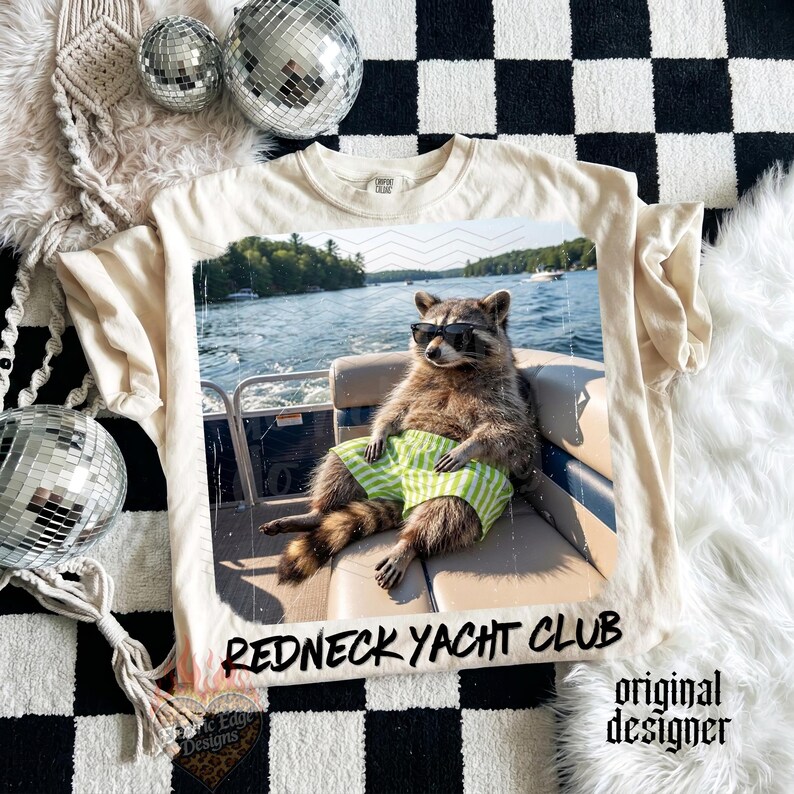 May include: Cream-colored t-shirt featuring a raccoon wearing sunglasses and green striped shorts, lounging on a boat. The image has the text "Redneck Yacht Club" and "original designer".