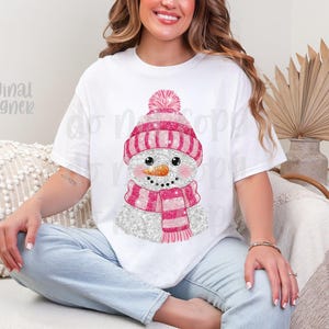 May include: White t-shirt with a glittery snowman design. The snowman has a pink and white striped hat and scarf, and a carrot nose. The text "original designer" is visible on the shirt. The shirt is worn by a person.