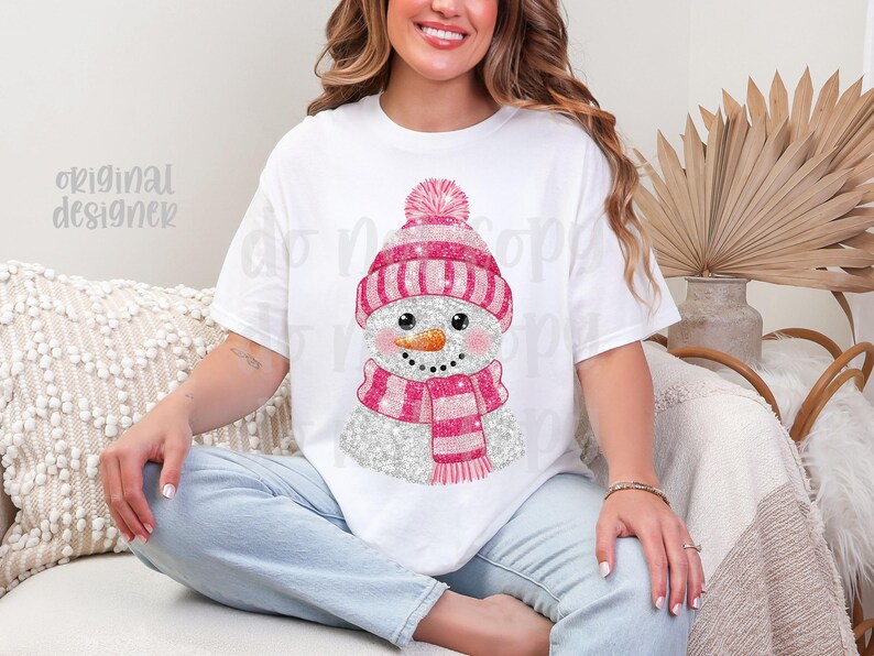 May include: White t-shirt with a glittery snowman design. The snowman has a pink and white striped hat and scarf, and a carrot nose. The text "original designer" is visible on the shirt. The shirt is worn by a person.