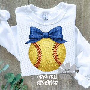 Pu&ograve; includere: Felpa bianca a maniche lunghe con un design a softball giallo, cuciture rosse e un fiocco blu. Le parole "original designer" sono stampate in basso. La felpa &egrave; su una superficie di legno.