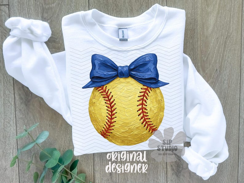 Pu&ograve; includere: Felpa bianca a maniche lunghe con un design a softball giallo, cuciture rosse e un fiocco blu. Le parole "original designer" sono stampate in basso. La felpa &egrave; su una superficie di legno.