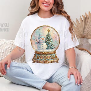 May include: A white short-sleeve t-shirt featuring a graphic print of a snow globe containing a snowman wearing a blue hat and scarf next to a decorated Christmas tree. The snow globe has an ornate gold base with intricate scrollwork details.