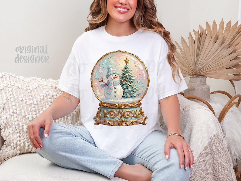 May include: A white short-sleeve t-shirt featuring a graphic print of a snow globe containing a snowman wearing a blue hat and scarf next to a decorated Christmas tree. The snow globe has an ornate gold base with intricate scrollwork details.