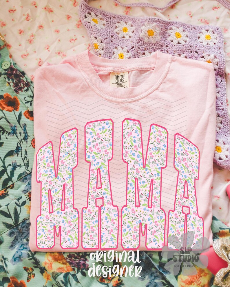 Op de afbeelding: Een lichtroze Comfort Colors sweatshirt met het woord "MAMA" in grote, bloemrijke letters met roze omtrek. De woorden "original designer" staan onderaan. Een gehaakte madeliefjesdeken is op de achtergrond.