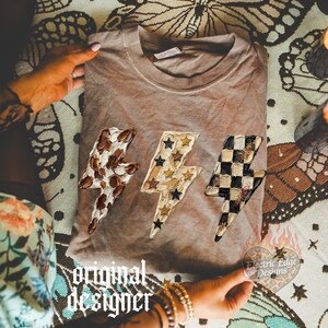 May include: A brown t-shirt featuring three lightning bolt designs with unique patterns: cow print, stars, and a checkerboard. The shirt is displayed on a patterned surface with the text "original designer" and "Electric Edge Designs".
