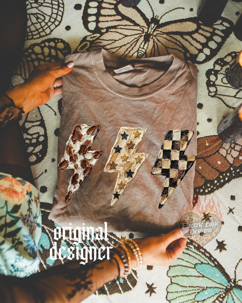 May include: A brown t-shirt featuring three lightning bolt designs with unique patterns: cow print, stars, and a checkerboard. The shirt is displayed on a patterned surface with the text "original designer" and "Electric Edge Designs".