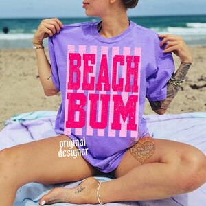 May include: A purple t-shirt with the words "BEACH BUM" in large, bright pink letters. The text is set against a striped pink and white background. The shirt is a casual, beach-themed apparel item.