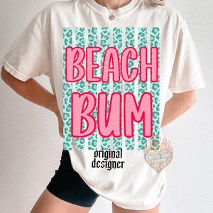May include: Cream-colored t-shirt with the words "BEACH BUM" in pink, outlined in red, over a teal and leopard print design. The shirt also features the text "original designer" in black.