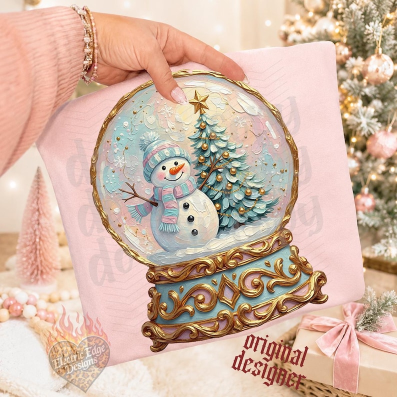 May include: A light pink sweatshirt featuring a graphic of a snow globe with a snowman wearing a blue and pink striped hat and scarf, standing next to a decorated Christmas tree with a gold star, set on an ornate gold and blue base.