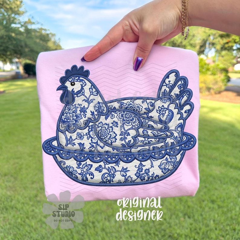 May include: A light pink sweatshirt featuring a blue and white floral design of a hen in a nest. The hen design is embroidered. The words "original designer" are printed in a script font.