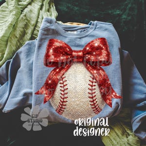 May include: A light blue sweatshirt featuring a baseball design. The baseball is embellished with sequins and red stitching, and a large red sequined bow is at the top. The words "original designer" are printed in white.