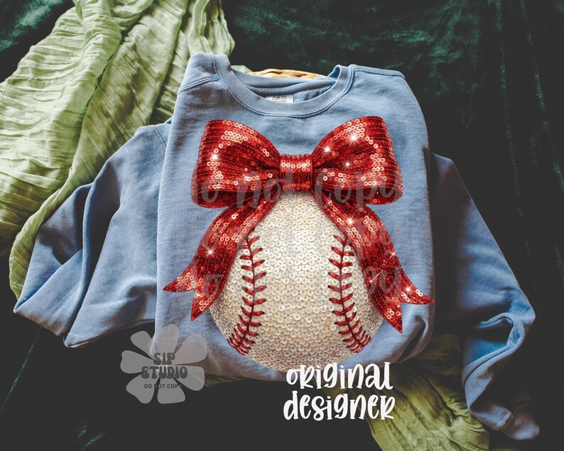 May include: A light blue sweatshirt featuring a baseball design. The baseball is embellished with sequins and red stitching, and a large red sequined bow is at the top. The words "original designer" are printed in white.