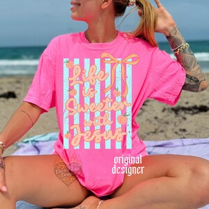 May include: A bright pink t-shirt with the text "Life is Sweeter with Jesus" in a playful font. The design includes peaches and a bow, set against blue and white stripes. The words "original designer" are printed on the bottom right.