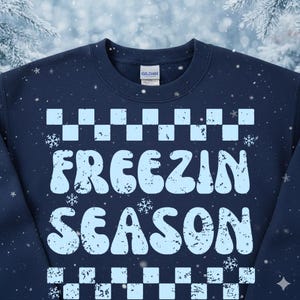 May include: Navy blue sweatshirt with the words "FREEZIN SEASON" in light blue, surrounded by a checkered pattern and snowflakes. The Gildan brand tag is visible at the back of the neck. A cozy winter-themed apparel item.
