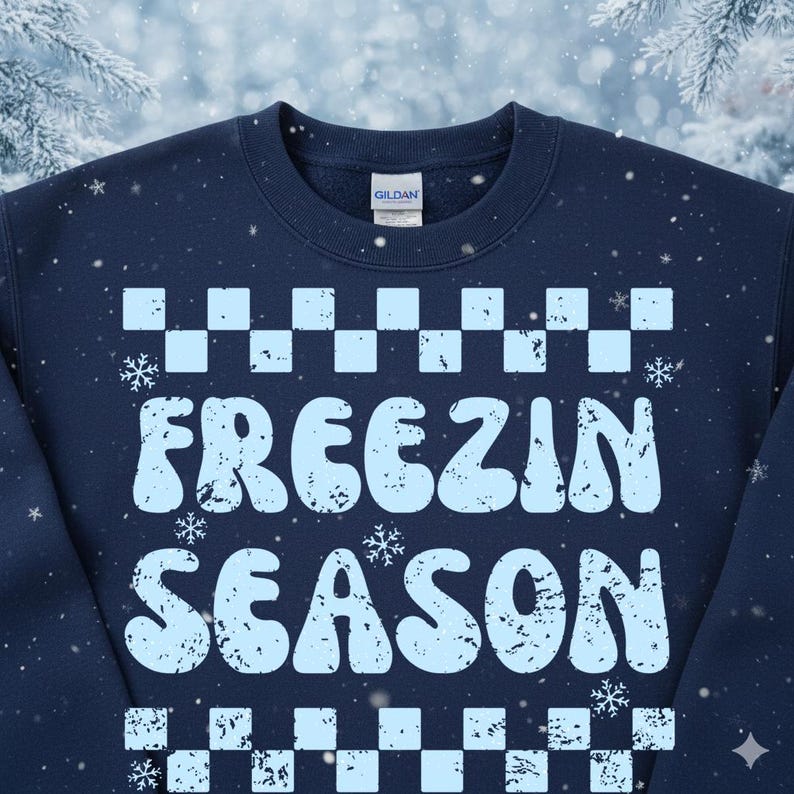 May include: Navy blue sweatshirt with the words "FREEZIN SEASON" in light blue, surrounded by a checkered pattern and snowflakes. The Gildan brand tag is visible at the back of the neck. A cozy winter-themed apparel item.