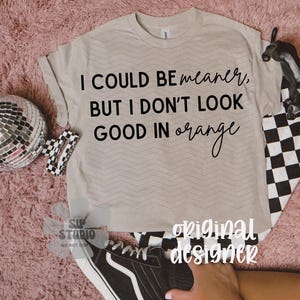 May include: A beige t-shirt with the text "I COULD BE meaner, BUT I DON'T LOOK GOOD IN orange" in black script. The shirt has a chevron pattern and is paired with a black and white checkered pattern and a black shoe.