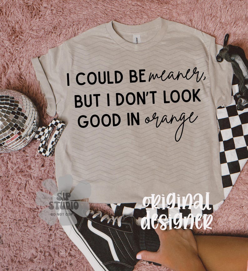 May include: A beige t-shirt with the text "I COULD BE meaner, BUT I DON'T LOOK GOOD IN orange" in black script. The shirt has a chevron pattern and is paired with a black and white checkered pattern and a black shoe.