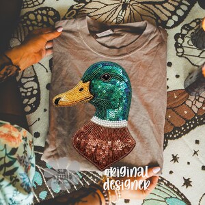 Pu&ograve; includere: Una maglietta color beige con un design dettagliato di un'anatra. La testa dell'anatra &egrave; verde con paillettes blu, un becco giallo perlato e un petto marrone con paillettes. Il testo "original designer" &egrave; visibile.