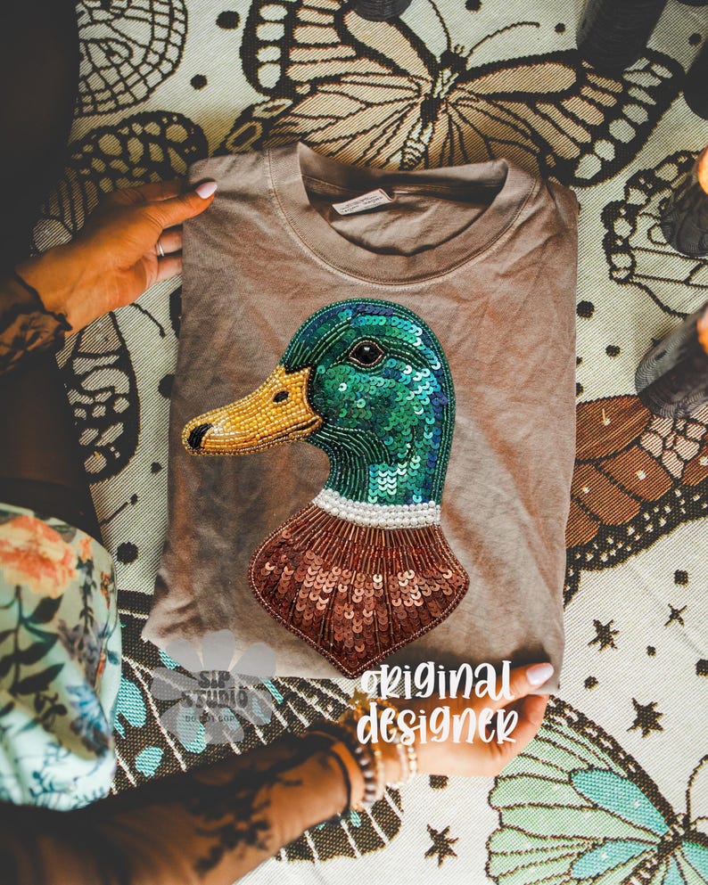 Pu&ograve; includere: Una maglietta color beige con un design dettagliato di un'anatra. La testa dell'anatra &egrave; verde con paillettes blu, un becco giallo perlato e un petto marrone con paillettes. Il testo "original designer" &egrave; visibile.