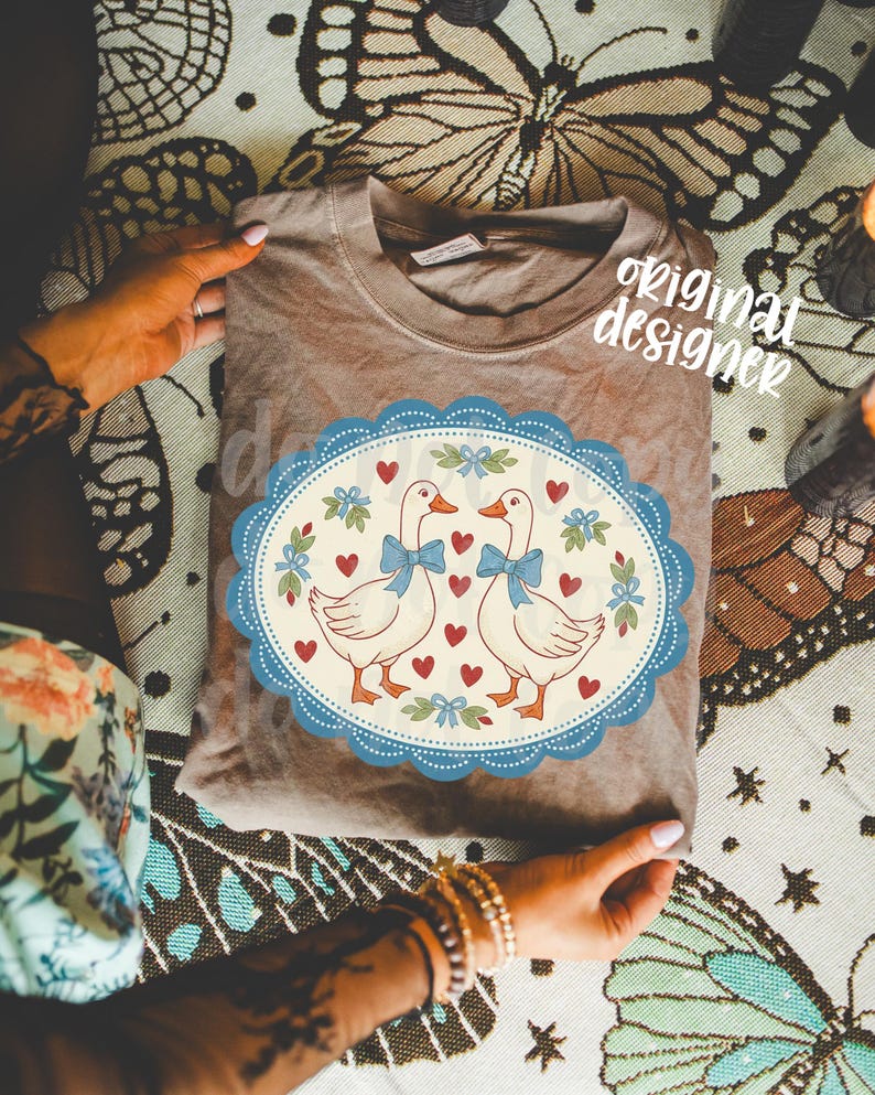 May include: A brown t-shirt featuring a whimsical design of two geese with blue bow ties, surrounded by hearts and floral accents within a blue scalloped oval. The words "original designer" are printed on the shirt.