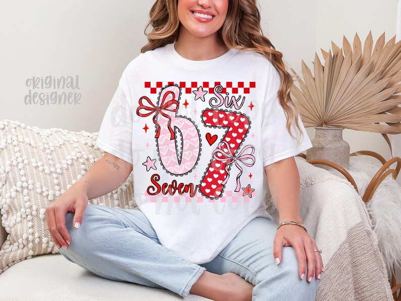 May include: White t-shirt with a colorful graphic design featuring the numbers "6" and "7" in a heart and polka dot pattern, with the words "Six" and "Seven". The design includes bows, stars, and a heart.