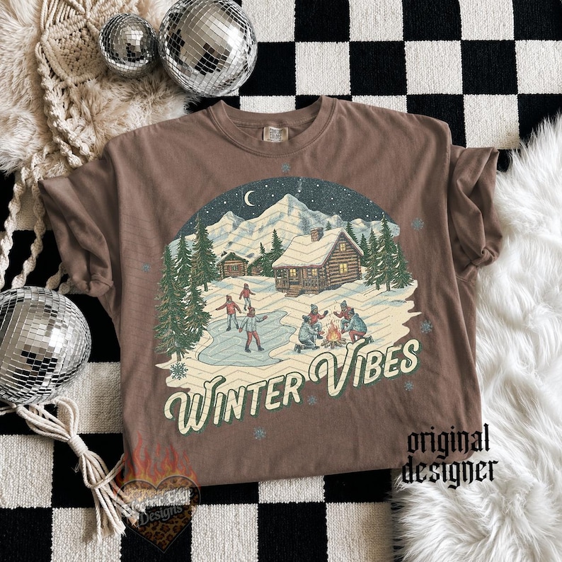 May include: A brown short-sleeve t-shirt featuring a graphic print of a snowy mountain landscape with a log cabin, pine trees, people ice skating, and a campfire. The text "WINTER VIBES" is printed in a cream-colored script font below the scene.