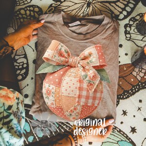 Op de afbeelding: Een bruin t-shirt met een patchwork perzikontwerp, een bloemenstrik en groene bladeren. De perzik is gemaakt van verschillende stoffen met patronen in roze, perzik en wit. De tekst "original designer" is zichtbaar.