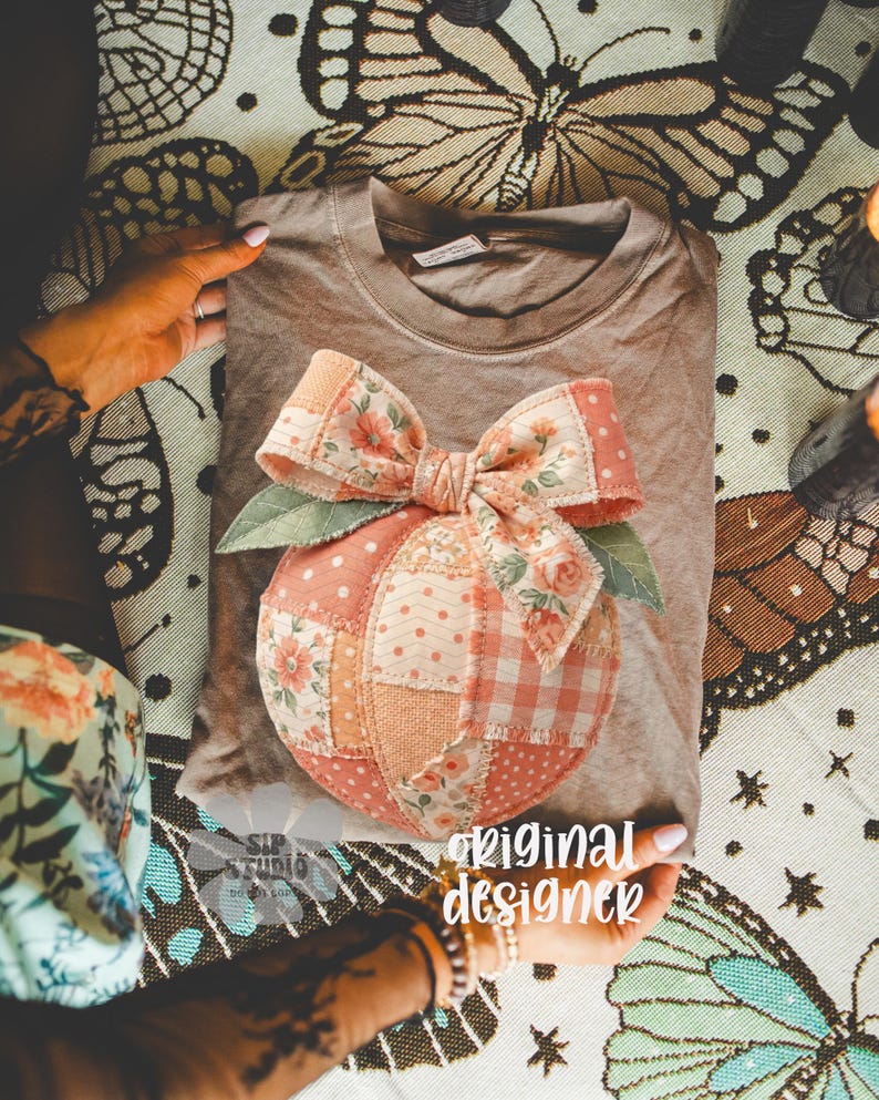 Op de afbeelding: Een bruin t-shirt met een patchwork perzikontwerp, een bloemenstrik en groene bladeren. De perzik is gemaakt van verschillende stoffen met patronen in roze, perzik en wit. De tekst "original designer" is zichtbaar.