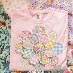 May include: A light pink t-shirt with a floral applique design made of various patterned fabrics. The shirt has a crew neck and the words "original designer" are printed on the bottom. A crocheted daisy-patterned item is visible in the background.