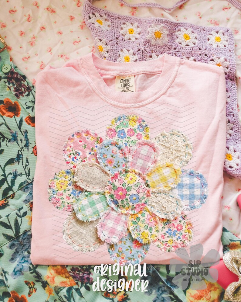 May include: A light pink t-shirt with a floral applique design made of various patterned fabrics. The shirt has a crew neck and the words "original designer" are printed on the bottom. A crocheted daisy-patterned item is visible in the background.