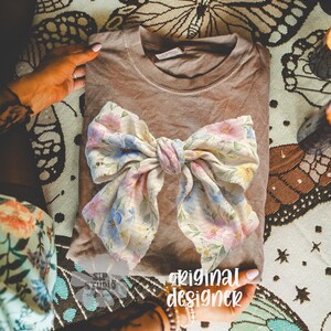 May include: A folded brown t-shirt with a large floral bow. The bow features a pattern of pink, blue, and yellow flowers on a cream background. The words "original designer" are visible in the image.