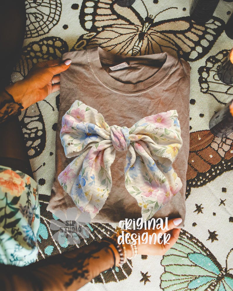 May include: A folded brown t-shirt with a large floral bow. The bow features a pattern of pink, blue, and yellow flowers on a cream background. The words "original designer" are visible in the image.