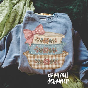 May include: A light blue sweatshirt featuring a stack of decorative bowls with floral and gingham patterns. A pink polka-dot bow is at the top. The words "original designer" are printed at the bottom.