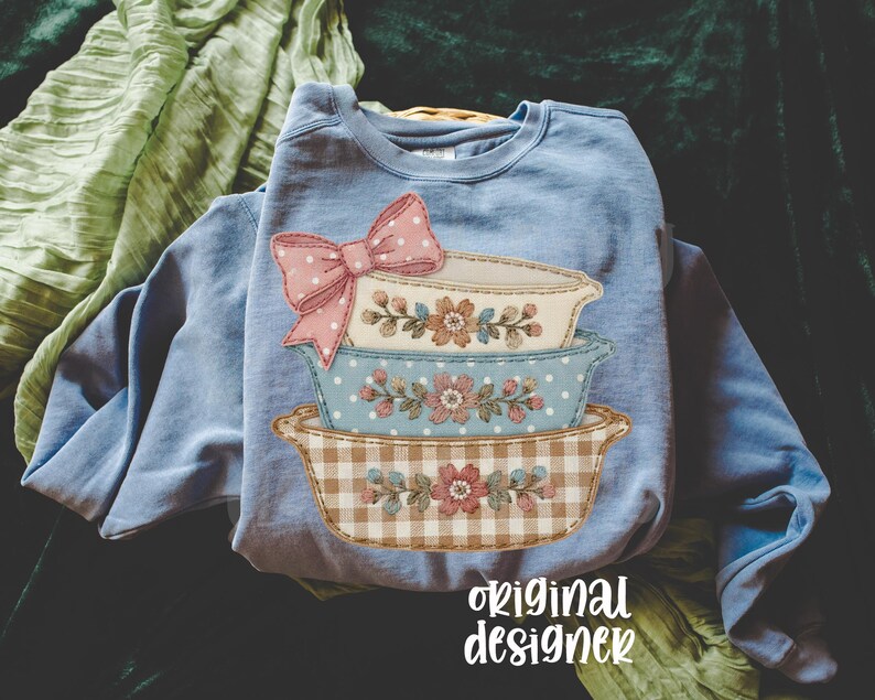 May include: A light blue sweatshirt featuring a stack of decorative bowls with floral and gingham patterns. A pink polka-dot bow is at the top. The words "original designer" are printed at the bottom.