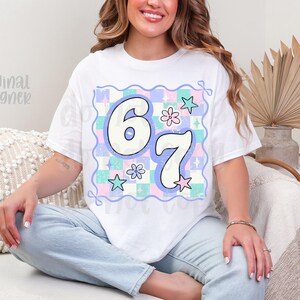 May include: White t-shirt featuring the numbers "67" in a playful, outlined font. The numbers are set against a pastel checkered background with stars and flowers. The shirt has a relaxed fit.