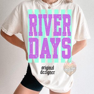 May include: Cream-colored t-shirt with the words "RIVER DAYS" in purple block letters over a teal striped background. The words "original designer" are printed below the graphic in black, along with a heart-shaped logo.