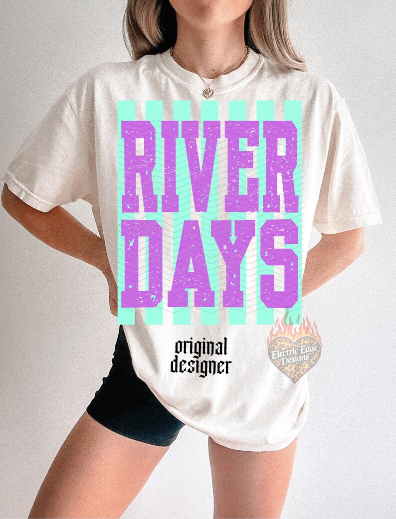 May include: Cream-colored t-shirt with the words "RIVER DAYS" in purple block letters over a teal striped background. The words "original designer" are printed below the graphic in black, along with a heart-shaped logo.