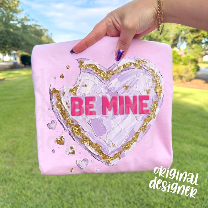 Op de afbeelding: Een lichtroze sweatshirt met een hartvormig ontwerp. Het hart heeft een getextureerde, geschilderde uitstraling met paarse en witte penseelstreken en is omlijnd met gouden glitter. De woorden "BE MINE" zijn roze. De woorden "original designer" zijn in het wit geschreven.