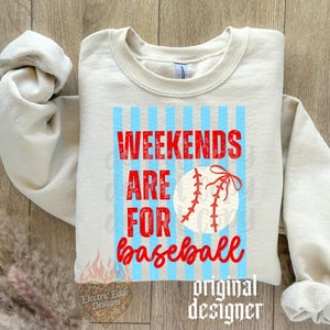 May include: A cream-coloured sweatshirt with the words "WEEKENDS ARE FOR baseball" in red script. The design includes a baseball graphic and light blue vertical stripes. The sweatshirt is a casual style.