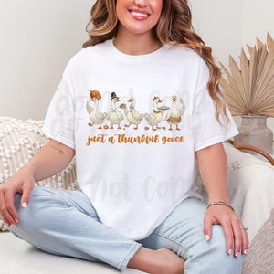 May include: White t-shirt featuring a graphic of five cartoon geese, each adorned with Thanksgiving-themed accessories like a pilgrim hat and a platter. The text "just a thankful goose" is printed in orange below the geese.