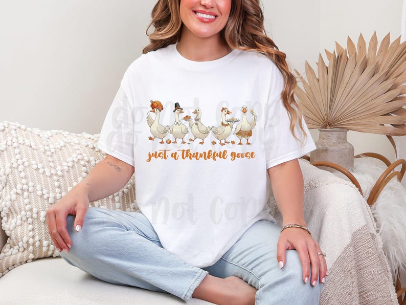 May include: White t-shirt featuring a graphic of five cartoon geese, each adorned with Thanksgiving-themed accessories like a pilgrim hat and a platter. The text "just a thankful goose" is printed in orange below the geese.