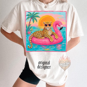 May include: Cream-colored t-shirt featuring a leopard wearing sunglasses, lounging on a pink flamingo float in a tropical scene. The design includes a sunset, palm tree, and the text "original designer".