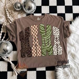 May include: A brown t-shirt featuring the word MAMA in large block letters. Each letter has a distinct pattern: leopard print, brown and white checkerboard, green zebra stripes, and black and white dalmatian spots. The text original designer is visible.