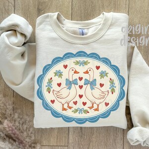 May include: A cream-colored sweatshirt featuring a whimsical design. The design includes two white geese with blue bows, surrounded by red hearts and floral accents within a blue scalloped oval. The words "original designer" are visible.