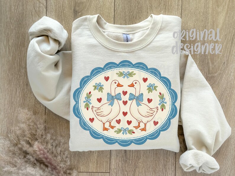May include: A cream-colored sweatshirt featuring a whimsical design. The design includes two white geese with blue bows, surrounded by red hearts and floral accents within a blue scalloped oval. The words "original designer" are visible.