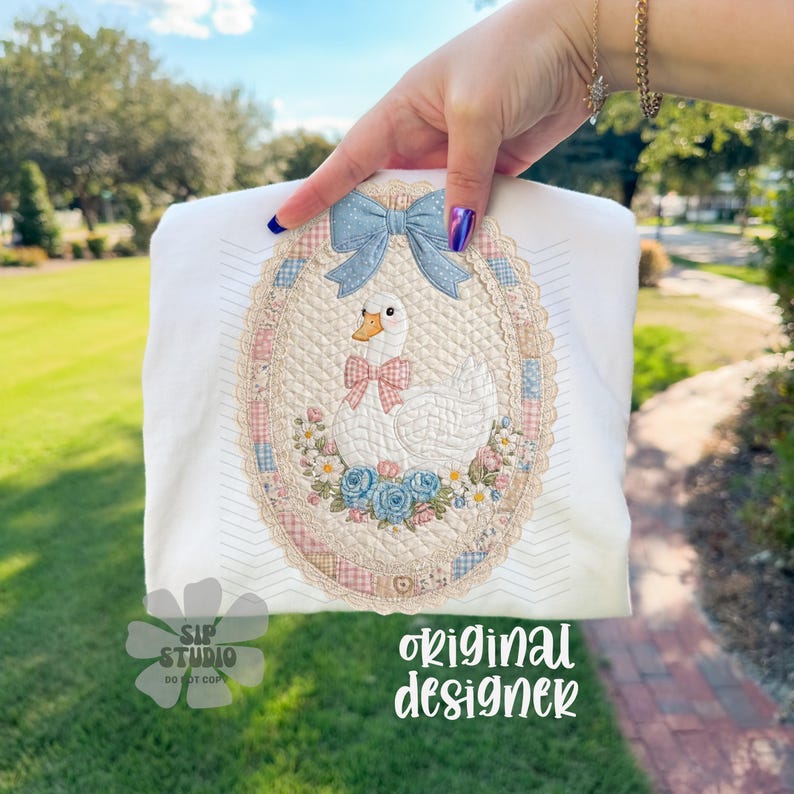 May include: White fabric with an embroidered oval design featuring a goose wearing a pink bow tie, surrounded by flowers and a blue bow. The design is framed by a lace-like border. The text "original designer" is visible.