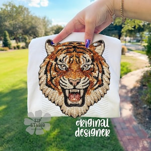 May include: White t-shirt featuring a detailed, embroidered tiger face with orange, brown, and gold sequins. The tiger's face is centered on the shirt, with the text "original designer" in a cursive font.