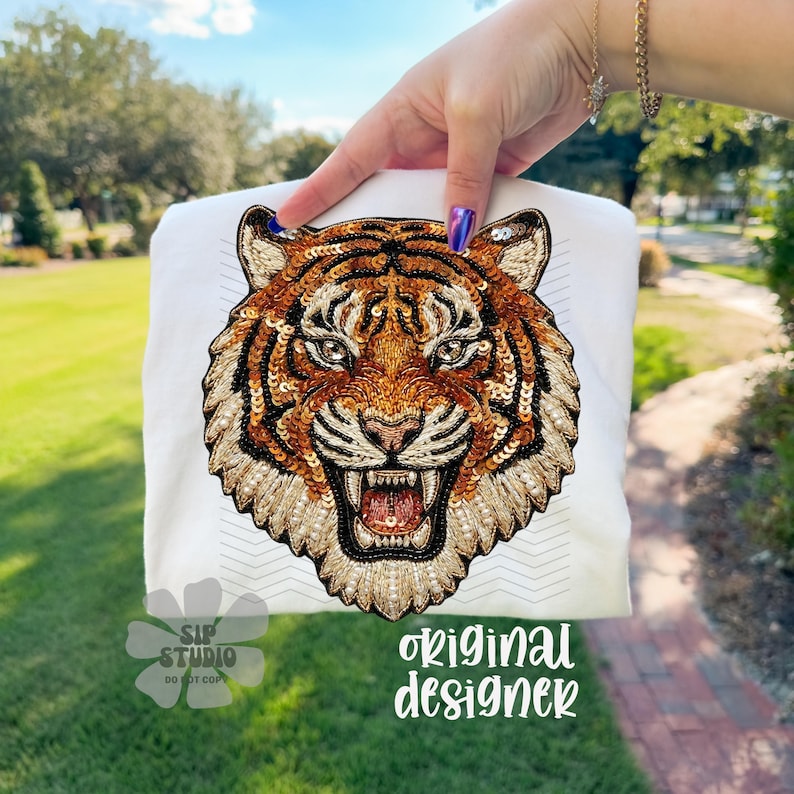 May include: White t-shirt featuring a detailed, embroidered tiger face with orange, brown, and gold sequins. The tiger's face is centered on the shirt, with the text "original designer" in a cursive font.